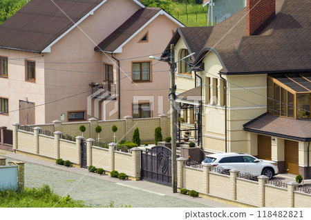 Quiet residential area with new expensive two- story cottages with brick fence and tile roofs and white sport car parked in front of garage door in paved well-kept yard. Luxury and prosperity concept. Quiet residential area with new expensive two- story cottages with brick fence and tile roofs and white sport car parked in front of garage door in paved well-kept yard. Luxury and prosperity concept. 118482821