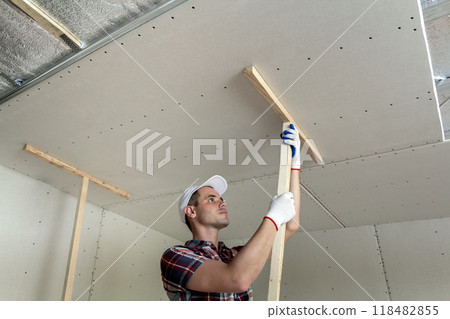 Young worker in protection work gloves fixing wooden holders for drywall suspended ceiling to metal frame insulated with shiny aluminum foil. DIY, do it yourself concept. 118482855