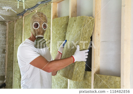 Young worker in gas mask insulating rock wool insulation staff in wooden frame for future house walls for cold and heat barrier. Comfortable warm home, economy, construction and renovation concept. 118482856