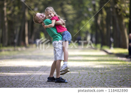 Two cute young funny smiling children, girl and boy, brother holding sister in his arms, having fun on blurred bright sunny park alley green trees bokeh background. Loving siblings relations concept. Two cute young funny smiling children, girl and boy, brother holding sister in his arms, having fun on blurred bright sunny park alley green trees bokeh background. Loving siblings relations concept. 118482890