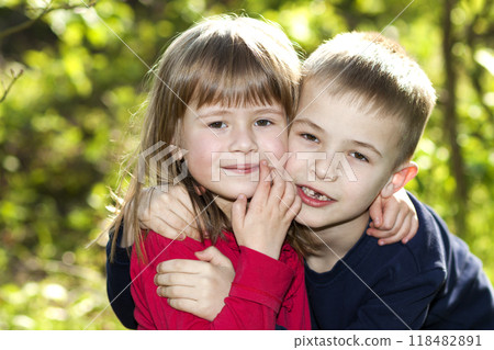 Two cute blond funny happy smiling children siblings, young boy brother embracing sister girl outdoors on bright sunny green bokeh background. Family relation, friendship and love concept. 118482891