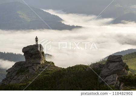 Silhouette of athletic climber tourist on high rocky formation on mountain valley filled with white puffy clouds and fog and covered with evergreen forest mountain slopes under clear sky background. 118482902