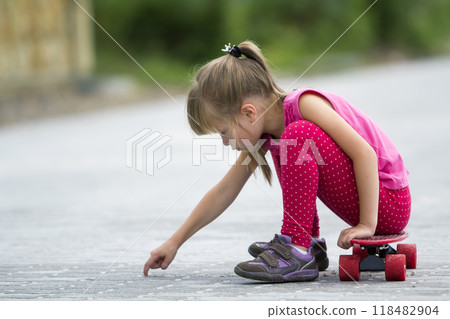 Pretty young long-haired blond child girl in casual pink clothing sitting on skateboard on paved suburb street on blurred sunny summer green background. Children activities, games and fun concept. Pretty young long-haired blond child girl in casual pink clothing sitting on skateboard on paved suburb street on blurred sunny summer green background. Children activities, games and fun concept. 118482904