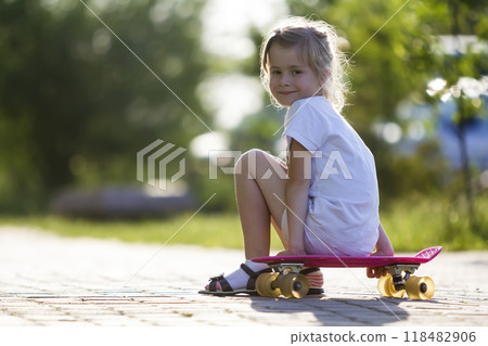 Pretty cute little blond girl in white shorts and T-shirt sitting on pink skateboard and smiling in camera on a light blurred summer background. Children activities and active lifestyle concept. 118482906