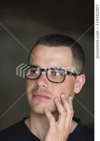Portrait of young handsome serious black-haired confident successful intellectual man in glasses with shiny black eyes looking thoughtfully into the space on dark background. 118482907