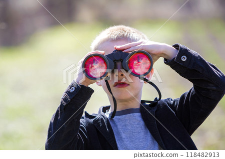 Portrait of little cute handsome cute blond boy watching intently something through binoculars in distance on blurred background. Children innocence, dreams, fantasies and imaginations concept. Portrait of little cute handsome cute blond boy watching intently something through binoculars in distance on blurred background. Children innocence, dreams, fantasies and imaginations concept. 118482913