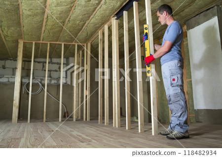 Interior of attic room with insulated ceiling and oak floor under reconstruction. Young professional worker uses level installing wooden frame for future walls. Renovation and improvement concept. Interior of attic room with insulated ceiling and oak floor under reconstruction. Young professional worker uses level installing wooden frame for future walls. Renovation and improvement concept. 118482934