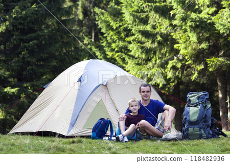 Holiday camping. Father and his son taking rest near tent after hiking in the forest Traveling and outdoors activities. Happy family relations and healthy lifestyle. 118482936