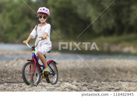 Cute young girl in white clothing, sunglasses with long braids wearing pink safety helmet riding child bicycle on pebbled river bank on blurred green summer background. Outdoors activity concept. 118482945