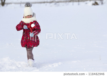 Cute little young funny toothless child girl in warm clothing playing having fun making snowballs on winter cold day on white bright blurred copy space background. Outdoors activity, holiday games. Cute little young funny toothless child girl in warm clothing playing having fun making snowballs on winter cold day on white bright blurred copy space background. Outdoors activity, holiday games. 118482946