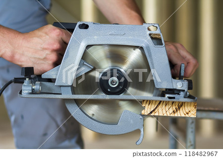 Close-up of muscular carpenter hands using new shiny modern powerful circular sharp electrical saw for cutting hard wooden board. Professional tools for construction and building concept. 118482967