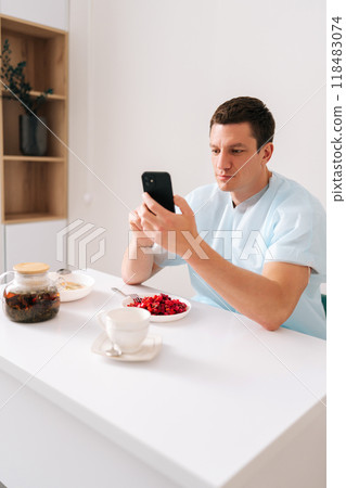 Vertical portrait of male patient in coat talking on video call using smartphone while sitting at table with meal in hospital room after medical procedure. Sick man resting recovering after surgery. 118483074