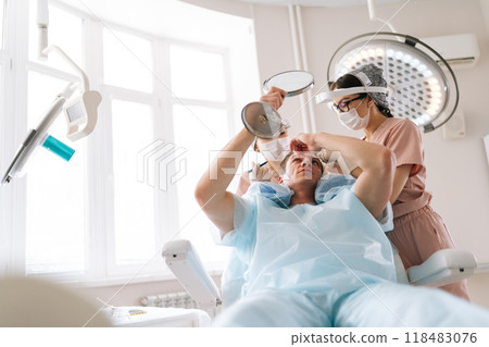Low-angle portrait of male patient with hairloss problem holding mirror, having consultation with trichologist, marking with pencil new hairline on head before hair transplantation surgery in clinic. 118483076