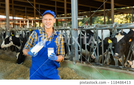 Smiling dairy farm owner pouring milk from bottle into glass in outdoor cowshed Smiling dairy farm owner pouring milk from bottle into glass in outdoor cowshed 118486413