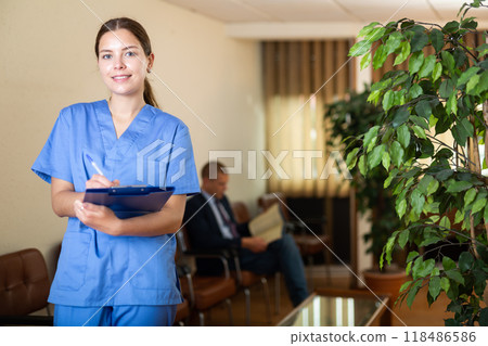 Positive young female doctor in clinic lobby, writes important notes Positive young female doctor in clinic lobby, writes important notes 118486586