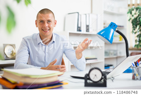 Smiling entrepreneur sitting at table in office, making welcome gesture Smiling entrepreneur sitting at table in office, making welcome gesture 118486748