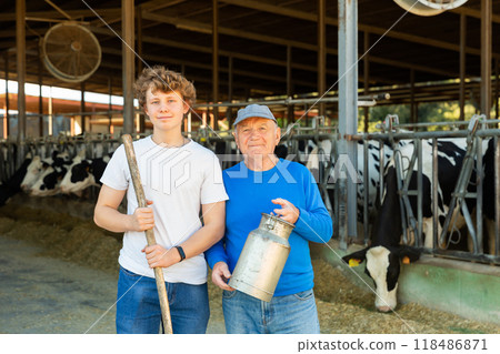 Elderly farmer and his assistant grandson with bottle of milk on the background of cows in stall 118486871