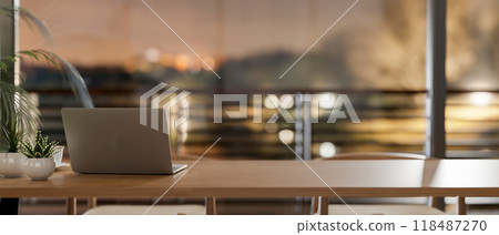 A laptop on a wooden tabletop in a room by the window, offering a dusk view through a balcony. 118487270