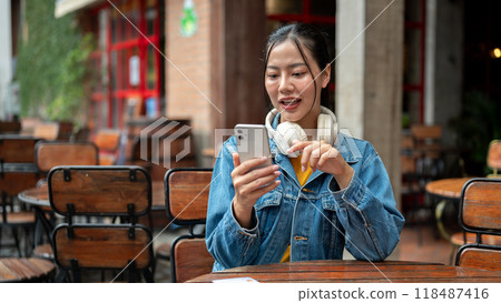 A positive Asian woman is using her smartphone while sitting at an outdoor table of a restaurant. 118487416