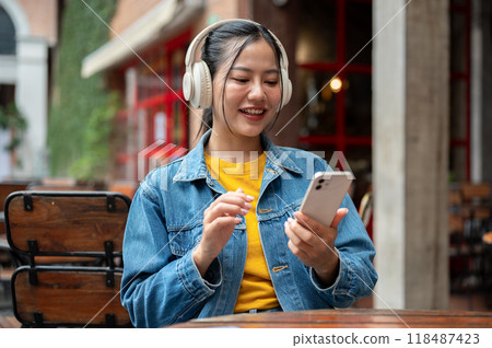 A happy Asian woman is using her phone and listening to music through her headphones at a restaurant 118487423