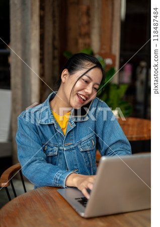 A woman is on the phone with her colleague while working on her laptop remotely from a coffee shop. 118487484