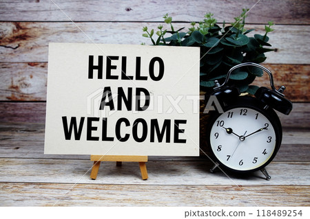 Hello and Welcome text on paper card and easel standing with alarm clock on wooden background 118489254