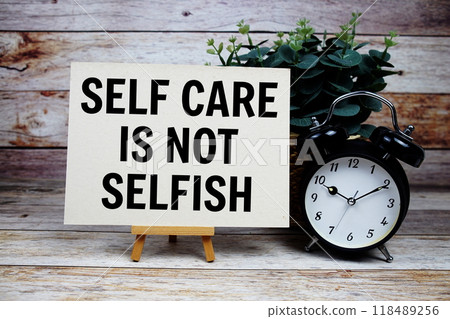 Self care is not selfish quotes text on paper card and easel standing with alarm clock on wooden background 118489256