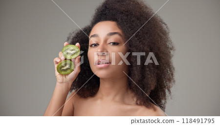 Beauty, vitamins and healthcare concept - beautiful African American woman with curly afro hairstyle and clean, healthy skin holding kiwi halves in her hands posing and looking at the camera Beauty, vitamins and healthcare concept - beautiful African American woman with curly afro hairstyle and clean, healthy skin holding kiwi halves in her hands posing and looking at the camera 118491795