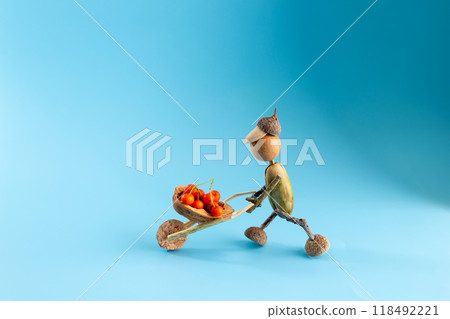 Charming autumn craft figurine made from acorns, with a body and head from acorns, twig limbs, pushing a twig wheelbarrow filled with red berries, blue gradient background, DIY kids activity, Charming autumn craft figurine made from acorns, with a body and head from acorns, twig limbs, pushing a twig wheelbarrow filled with red berries, blue gradient background, DIY kids activity, 118492221