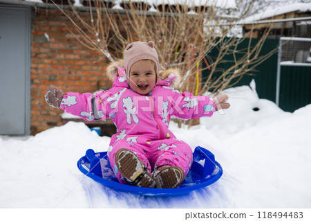 A child sledding down a snowy hill in a winter backyard. A happy child laughs and slides down an icy hill on a winter day. Perfect for capturing a child, a sled, and winter. 118494483