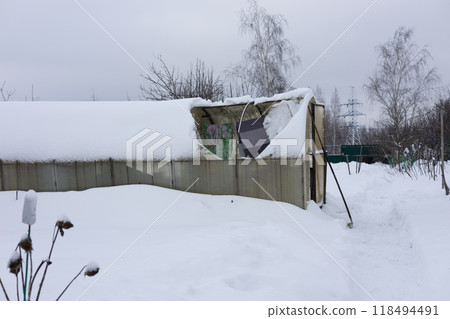 An old greenhouse with a collapsed roof, covered in snow, in a winter garden, demonstrating the destructive power of nature. During a heavy snowfall in winter, the roof of the old greenhouse broke. 118494491