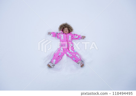 A small child makes a snow angel, dressed in a pink snowsuit, lying on the white fluffy snow. The child lies on his back in the fresh snow, arms and legs spread out. Childhood in winter outside. A small child makes a snow angel, dressed in a pink snowsuit, lying on the white fluffy snow. The child lies on his back in the fresh snow, arms and legs spread out. Childhood in winter outside. 118494495
