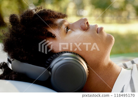 Profile shot of teenage African American boy lying on green grass in park while listening to music through headphones lost in thoughts and imagination 118495170