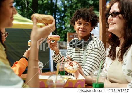 Medium shot of smiling teenage Black boy holding delicious donut sitting at cafe table with friends while enjoying spending time together outside in sunshine, copy space Medium shot of smiling teenage Black boy holding delicious donut sitting at cafe table with friends while enjoying spending time together outside in sunshine, copy space 118495267