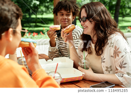 Side view of smiling teenage girl sitting with friends at cafe table celebrating birthday with delicious donuts outside in city park Side view of smiling teenage girl sitting with friends at cafe table celebrating birthday with delicious donuts outside in city park 118495285