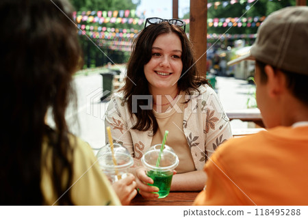 Portrait shot of charming teenage girl sitting at cafe table in park listening with smile to friends while enjoying time together and summer day outside, copy space 118495288
