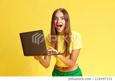 Young attractive girl expressing shock, surprise while using laptop and making order at online shopping against bright yellow background. 118497131