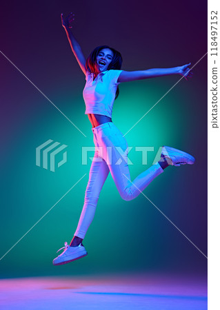 Full length photo of attractive young girl, jumping of success raising hands and shouting in neon light against gradient studio background. 118497152