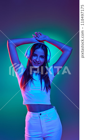 Dressed casual white clothes, dancing listening music in headphones in neon light against gradient blue-green background. Dressed casual white clothes, dancing listening music in headphones in neon light against gradient blue-green background. 118497175