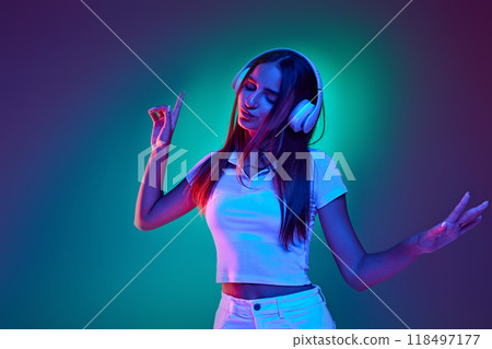 Dynamic photo of woman dressed casual attire and headphones listening music and singing in neon light against gradient studio background. Dynamic photo of woman dressed casual attire and headphones listening music and singing in neon light against gradient studio background. 118497177