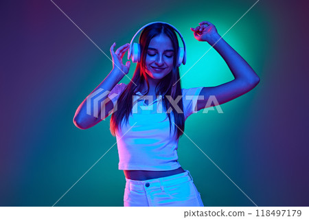Young beautiful girl, listening music in headphones and dancing raising hands in neon light against gradient studio background. 118497179