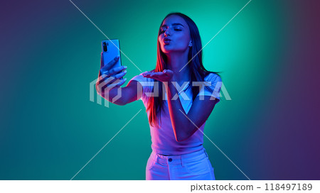 Portrait of young girl, sending air-kiss her friend while talking with friend online on phone in neon light against white studio background. Portrait of young girl, sending air-kiss her friend while talking with friend online on phone in neon light against white studio background. 118497189