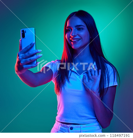 Young charming girl talking online using phone and waving hello in neon light against gradient studio background. Young charming girl talking online using phone and waving hello in neon light against gradient studio background. 118497191