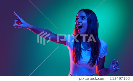 Young girl dressed casual attire looking at distance and pointing in neon light against gradient studio background. Start of sales season 118497193