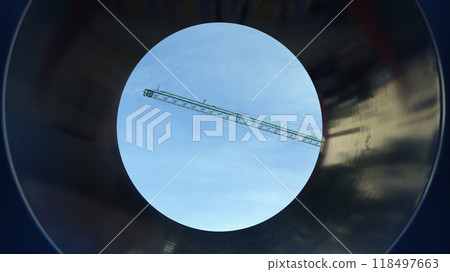 Looking through a pipe at a construction site, with a crane and clear blue sky visible above the industrial area 118497663
