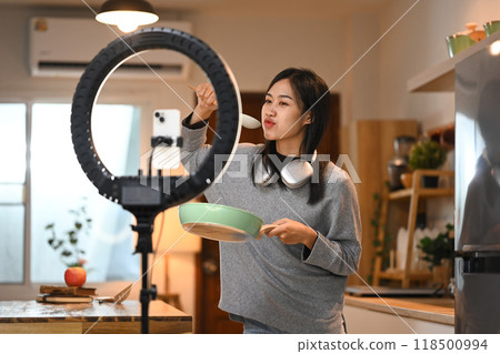 Cheerful young female food blogger recording video in kitchen with a smartphone on a tripod stand with a ring light 118500994