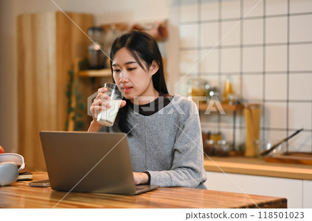Pretty young asian woman drinking milk while using a laptop at wooden table in a kitchen 118501023