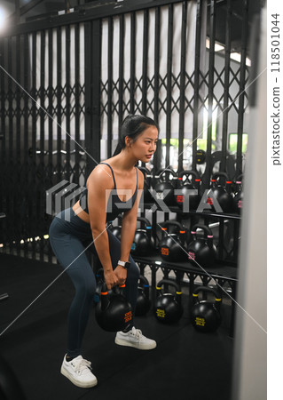Focused young beautiful woman exercising with kettle bell in a gym. Sports and healthy lifestyle concept Focused young beautiful woman exercising with kettle bell in a gym. Sports and healthy lifestyle concept 118501044