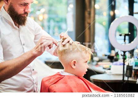 Man barber cutting little boy's hair using comb and scissors. Child getting haircut from adult male, likely barber. Professional hairdresser and cute client at modern barbershop. Man barber cutting little boy's hair using comb and scissors. Child getting haircut from adult male, likely barber. Professional hairdresser and cute client at modern barbershop. 118501082