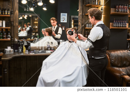Experienced hairdresser styling client's hair. Handsome hairstylist in fancy outfit at work. Male barber using professional hairdryer and brush. 118501101